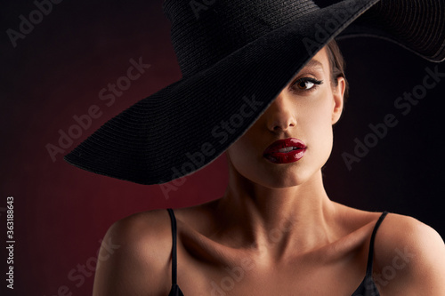 portrait of beautiful tanned girl with professional makeup, red lips, on a burgundy background in a black dress with straps and black hat that covers her eye, she looks at the camera with serious face © monchak