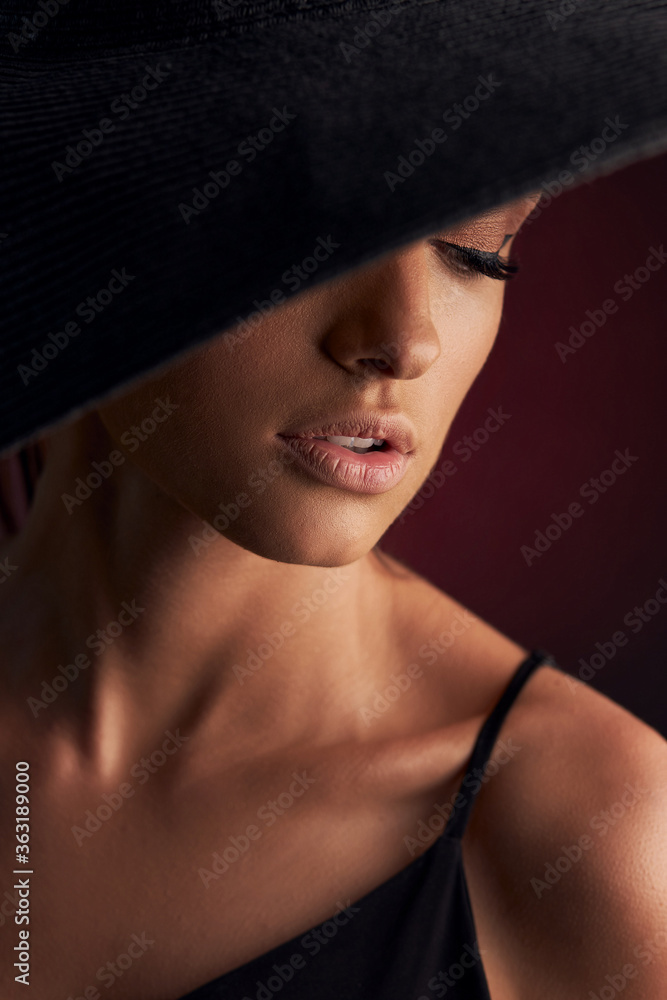 portrait of a beautiful tanned girl with professional makeup on a burgundy background in a black dress with straps and black hat that covers her eye portrait of a beautiful tanned girl with professional makeup on a burgundy background in a black dress with straps and black hat that covers her eye