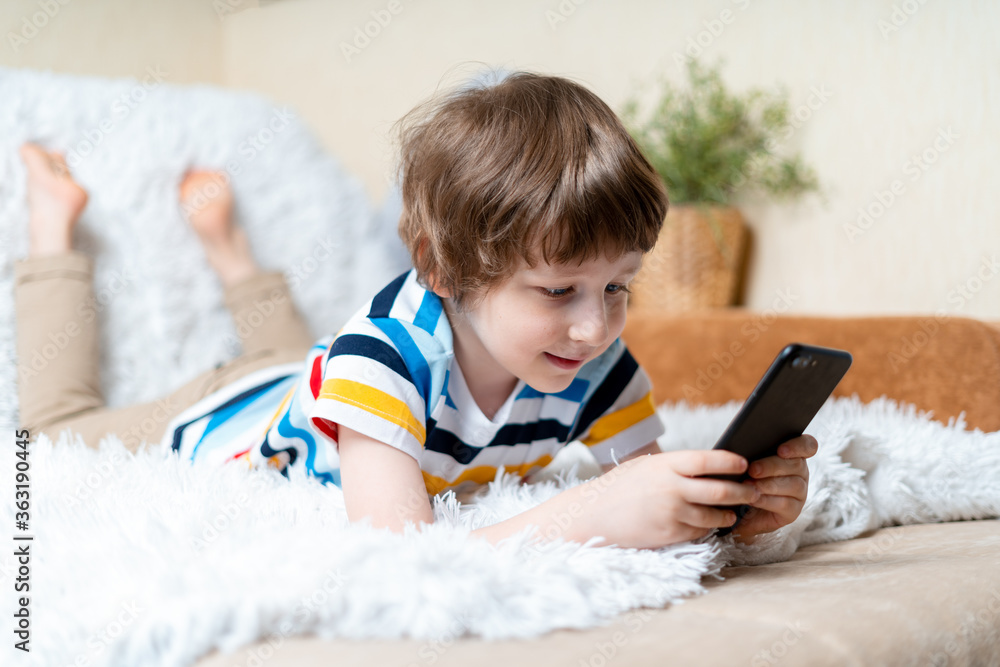 Happy little child boy playing online game, watching video on cellphone, lying on sofa entertaining in living room. Smiling small kid using funny mobile apps, enjoying free leisure time at home. Happy little child boy playing online game, watching video on cellphone, lying on sofa entertaining in living room. Smiling small kid using funny mobile apps, enjoying free leisure time at home.