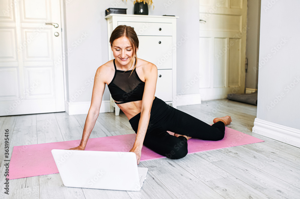 An online attractive female coach is talking to a client using a webcamera before online workout. She sits on a fitness mat at home wearing black sportswear An online attractive female coach is talking to a client using a webcamera before online workout. She sits on a fitness mat at home wearing black sportswear