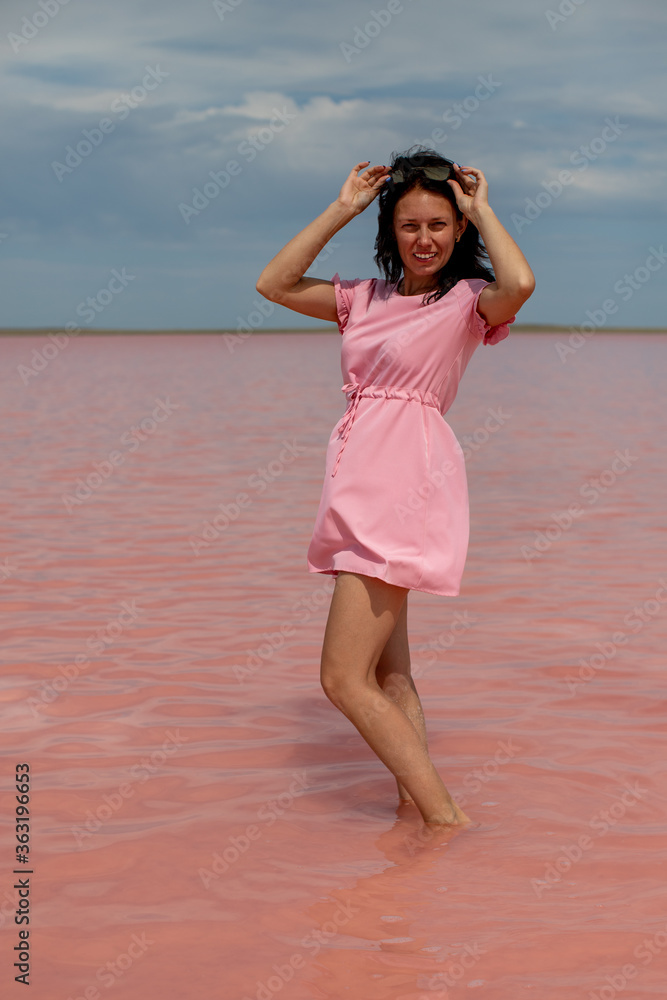 Brunette in the pink dress puts on sunglasses and posing against the background of the pink Lake Kobeituz in north Kazakhstan's Akmola Region. Brunette in the pink dress puts on sunglasses and posing against the background of the pink Lake Kobeituz in north Kazakhstan's Akmola Region.