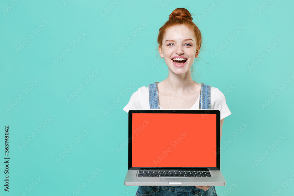 Naklejka premium Cheerful young readhead girl in casual denim clothes posing isolated on blue turquoise background studio. People lifestyle concept. Mock up copy space. Hold laptop pc computer with blank empty screen.