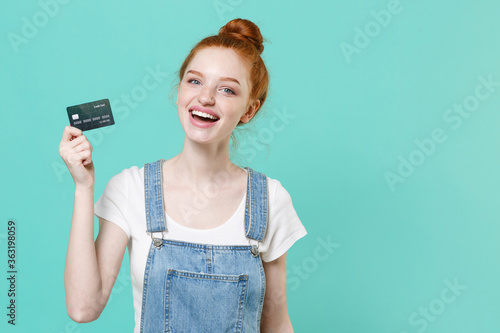Cheerful young readhead girl in casual denim clothes isolated on blue turquoise wall background studio portrait. People sincere emotions lifestyle concept. Mock up copy space. Hold credit bank card.
