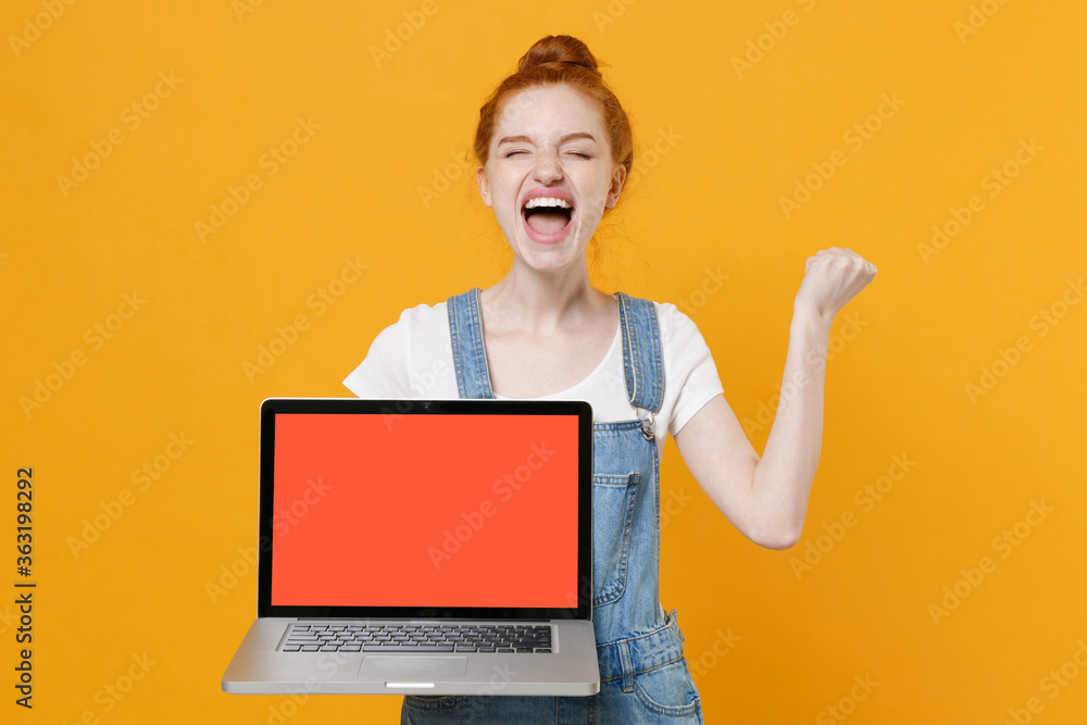 Screaming young girl in denim clothes white t-shirt isolated on yellow background. People lifestyle concept. Hold laptop pc computer with blank empty screen doing winner gesture keeping eyes closed. Screaming young girl in denim clothes white t-shirt isolated on yellow background. People lifestyle concept. Hold laptop pc computer with blank empty screen doing winner gesture keeping eyes closed.
