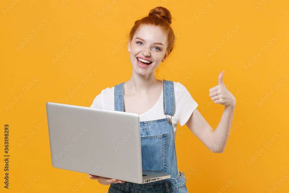 Funny young readhead girl in casual denim clothes white t-shirt isolated on yellow background studio. People lifestyle concept. Mock up copy space. Working on laptop pc computer, showing thumb up. Funny young readhead girl in casual denim clothes white t-shirt isolated on yellow background studio. People lifestyle concept. Mock up copy space. Working on laptop pc computer, showing thumb up.