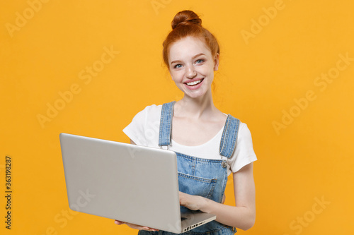 Smiling young readhead girl in casual denim clothes white t-shirt isolated on yellow background studio portrait. People emotions lifestyle concept. Mock up copy space. Working on laptop pc computer.