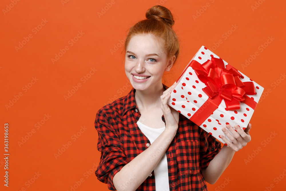 Smiling young readhead girl in checkered shirt posing isolated on orange background. St. Valentine's Day International Women's Day birthday holiday concept. Hold red present box with gift ribbon bow. Smiling young readhead girl in checkered shirt posing isolated on orange background. St. Valentine's Day International Women's Day birthday holiday concept. Hold red present box with gift ribbon bow.