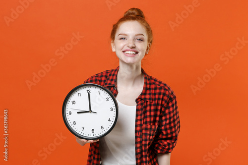 Smiling young readhead girl in casual red checkered shirt posing isolated on orange wall background studio portrait. People sincere emotions lifestyle concept. Mock up copy space. Hold in hand clock.