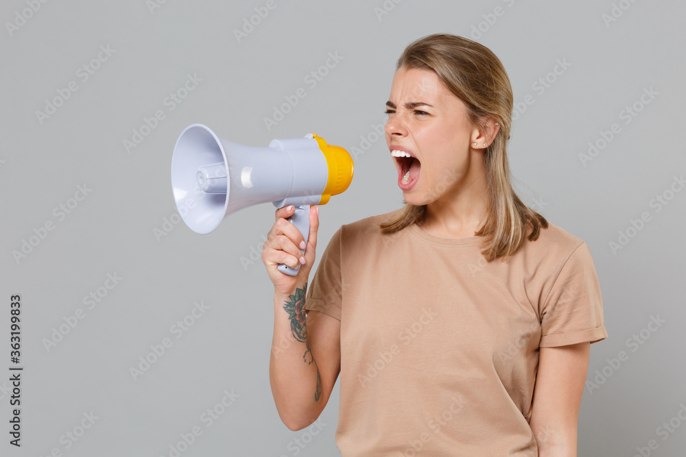 Displeased young blonde woman girl in casual beige t-shirt posing isolated on gray wall background studio portrait. People lifestyle concept. Mock up copy space. Screaming in megaphone, looking aside. Displeased young blonde woman girl in casual beige t-shirt posing isolated on gray wall background studio portrait. People lifestyle concept. Mock up copy space. Screaming in megaphone, looking aside.
