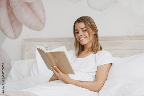 Smiling pretty young woman girl in white t-shirt lying in bed with white sheet pillow blanket spending time in bedroom at home. Rest relax good mood lifestyle concept. Mock up copy space. Read book.