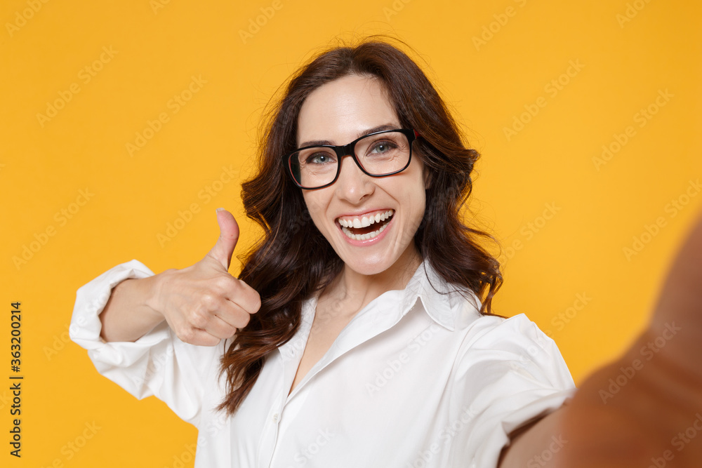Close up of cheerful young brunette business woman in white shirt glasses isolated on yellow background. Achievement career wealth business concept. Doing selfie shot on mobile phone showing thumb up. Close up of cheerful young brunette business woman in white shirt glasses isolated on yellow background. Achievement career wealth business concept. Doing selfie shot on mobile phone showing thumb up.
