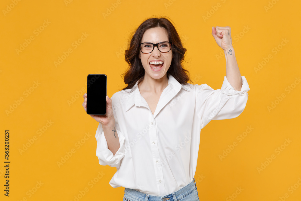 Joyful young brunette business woman in white shirt glasses isolated on yellow background. Achievement career wealth business concept. Hold mobile phone with blank empty screen doing winner gesture. Joyful young brunette business woman in white shirt glasses isolated on yellow background. Achievement career wealth business concept. Hold mobile phone with blank empty screen doing winner gesture.
