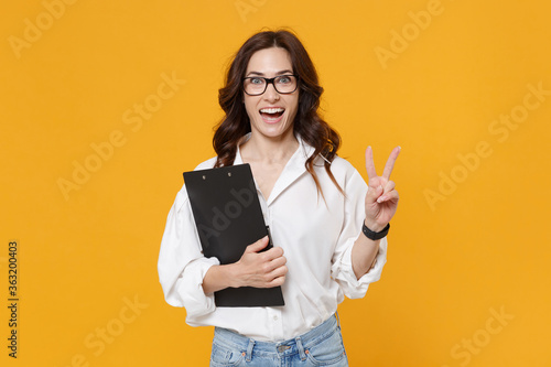 Excited business woman in white shirt glasses isolated on yellow background. Achievement career wealth business concept. Mock up copy space. Hold clipboard with papers document, showing victory sign.