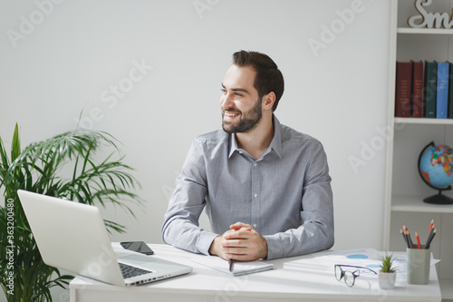 Smiling handsome young bearded business man in gray shirt sitting at desk, work on laptop pc computer in light office on white wall background. Achievement business career concept. Looking aside.