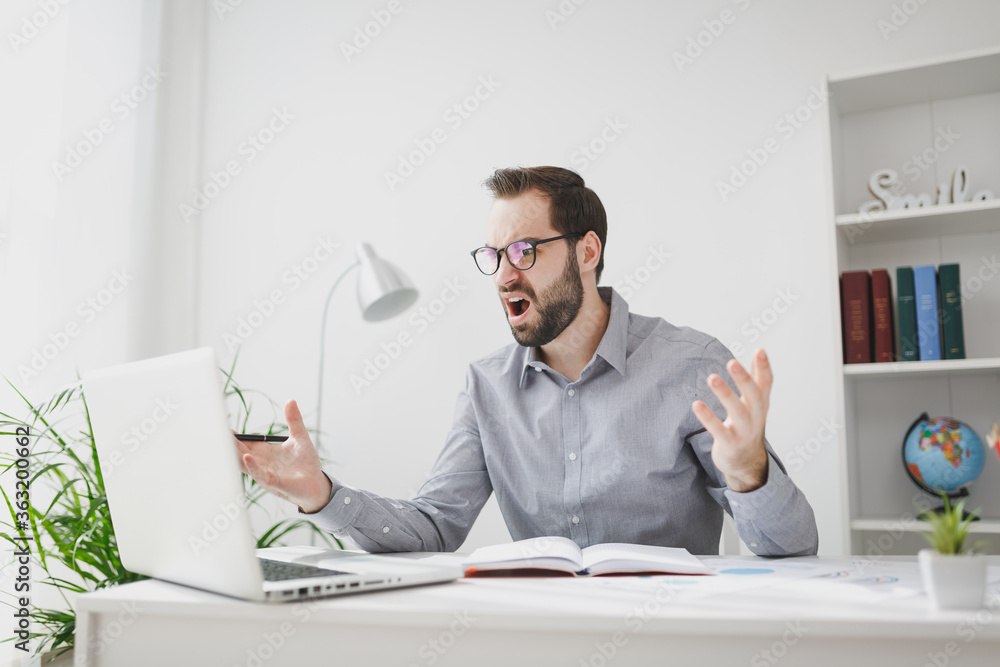 Angry young bearded business man in gray shirt glasses sit at desk in light office on white wall background. Achievement business career concept. Work on laptop pc computer hold pen spreading hands. Angry young bearded business man in gray shirt glasses sit at desk in light office on white wall background. Achievement business career concept. Work on laptop pc computer hold pen spreading hands.