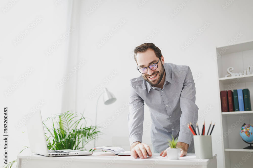 Smiling young bearded business man in gray shirt standing near desk working with laptop computer papers document notebook in light office on white wall background. Achievement business career concept. Smiling young bearded business man in gray shirt standing near desk working with laptop computer papers document notebook in light office on white wall background. Achievement business career concept.