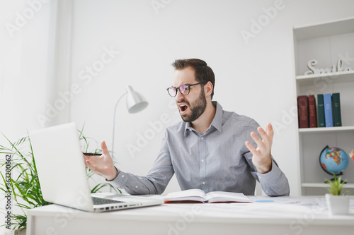 Angry young bearded business man in gray shirt glasses sit at desk in light office on white wall background. Achievement business career concept. Work on laptop pc computer hold pen spreading hands.