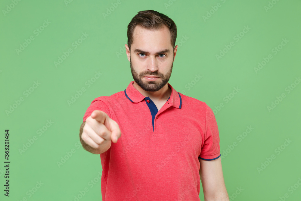 Strict young bearded man guy in casual red pink t-shirt posing isolated on green background studio portrait. People emotions lifestyle concept. Mock up copy space. Pointing index finger on camera. Strict young bearded man guy in casual red pink t-shirt posing isolated on green background studio portrait. People emotions lifestyle concept. Mock up copy space. Pointing index finger on camera.
