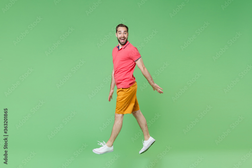 Side view of excited young bearded man guy in casual red pink t-shirt posing isolated on green wall background studio portrait. People lifestyle concept. Mock up copy space. Jumping, looking camera. Side view of excited young bearded man guy in casual red pink t-shirt posing isolated on green wall background studio portrait. People lifestyle concept. Mock up copy space. Jumping, looking camera.