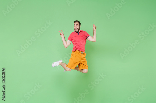 Excited young bearded man guy in casual red pink t-shirt posing isolated on green wall background studio portrait. People emotions lifestyle concept. Mock up copy space. Jumping, showing victory sign.
