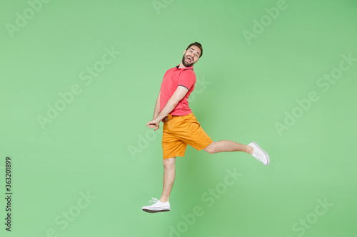 Side view of funny young bearded man guy in casual red pink t-shirt posing isolated on green background studio portrait. People lifestyle concept. Mock up copy space. Jumping, dancing, spreading legs. © ViDi Studio