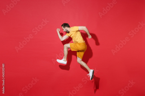 Side view of screaming young bearded man guy in casual yellow t-shirt posing isolated on red background studio portrait. People emotions lifestyle concept. Mock up copy space. Jumping like running.