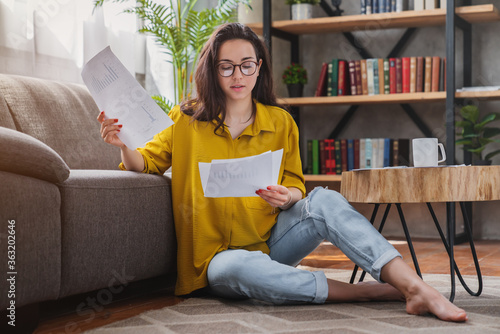 Fényképezés Young woman looking at bills while sitting on floor at living room managing home