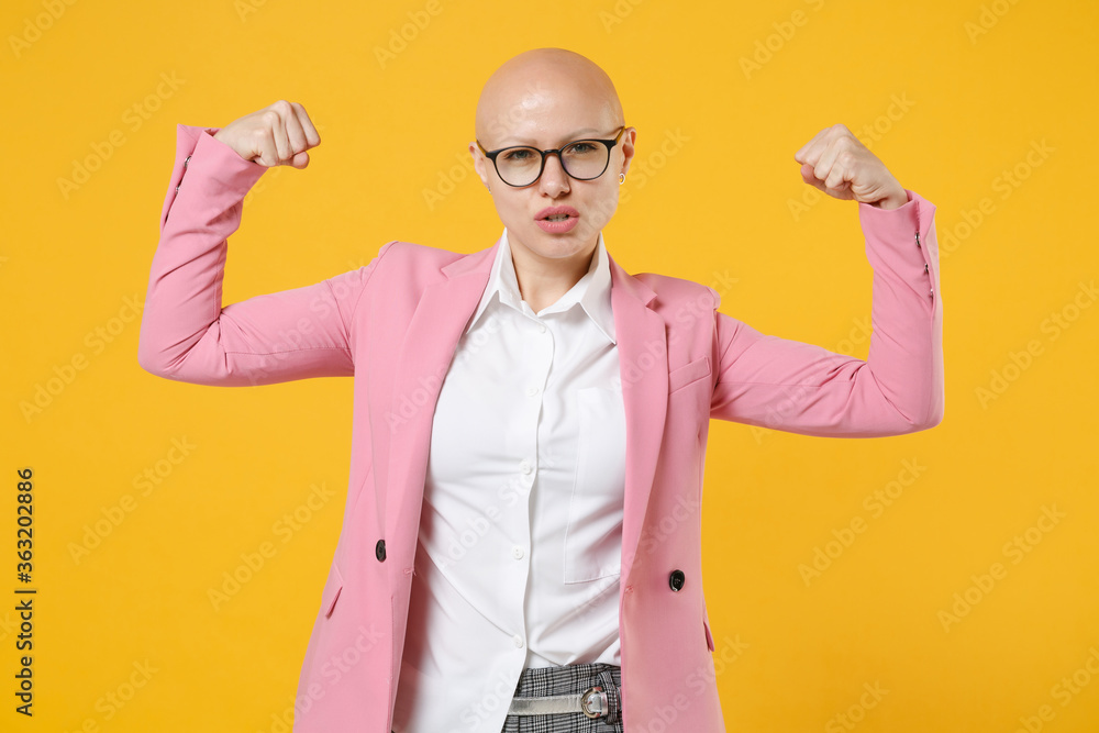 Strong young bald business woman in white shirt pink jacket glasses isolated on yellow wall background studio. Achievement career wealth business concept. Mock up copy space. Showing biceps, muscles. Strong young bald business woman in white shirt pink jacket glasses isolated on yellow wall background studio. Achievement career wealth business concept. Mock up copy space. Showing biceps, muscles.