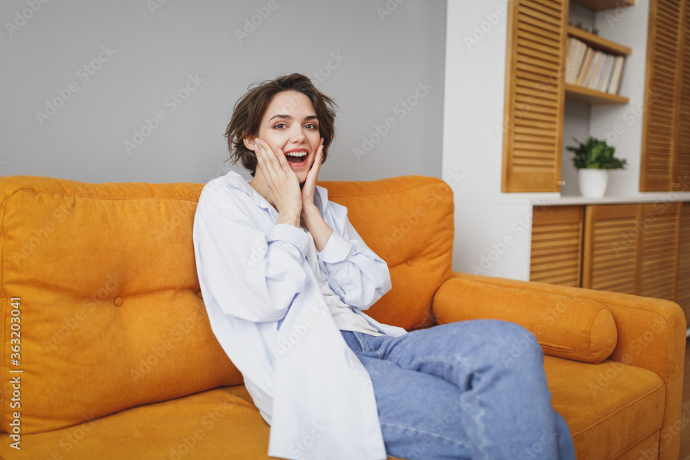 Amazed shocked young brunette woman girl in casual clothes sit on couch spending time in living room at home. Rest relax good mood leisure lifestyle concept. Mock up copy space. Put hands on cheeks. Amazed shocked young brunette woman girl in casual clothes sit on couch spending time in living room at home. Rest relax good mood leisure lifestyle concept. Mock up copy space. Put hands on cheeks.