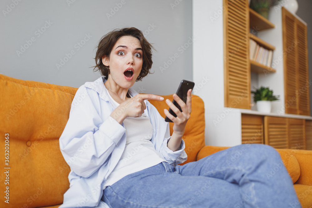 Shocked young woman girl in casual clothes sit on couch spending time in living room at home. Rest relax good mood leisure lifestyle concept. Mock up copy space. Pointing index finger on mobile phone.