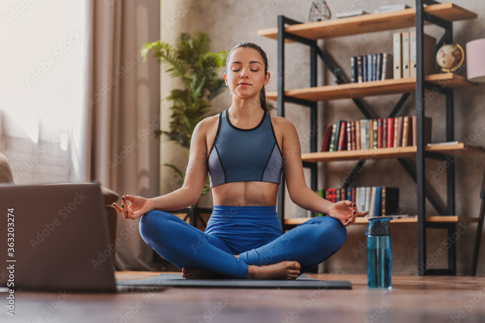 Online yoga maditation home. Young girl sitting in lotus pose near laptop at home Online yoga maditation home. Young girl sitting in lotus pose near laptop at home