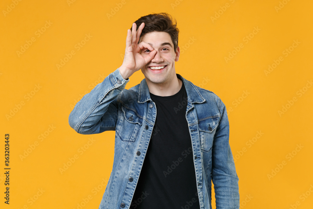 Smiling young man guy wearing casual denim jacket posing isolated on yellow background in studio. People lifestyle concept. Mock up copy space. Holding hand near eyes, imitating glasses or binoculars. Smiling young man guy wearing casual denim jacket posing isolated on yellow background in studio. People lifestyle concept. Mock up copy space. Holding hand near eyes, imitating glasses or binoculars.