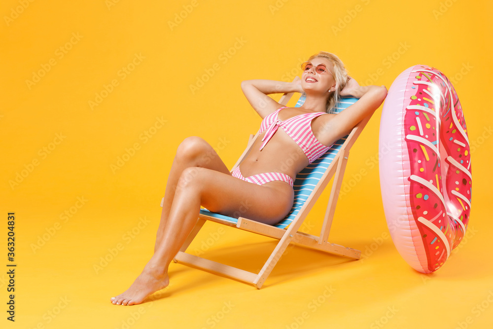 Smiling young woman girl in pink striped swimsuit sit on deck chair isolated on yellow background studio. People summer vacation rest lifestyle concept. Mock up copy space. Holding hands behind head. Smiling young woman girl in pink striped swimsuit sit on deck chair isolated on yellow background studio. People summer vacation rest lifestyle concept. Mock up copy space. Holding hands behind head.