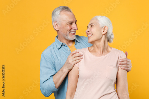 Smiling elderly gray-haired couple woman man in casual clothes posing isolated on yellow wall background studio portrait. People lifestyle concept. Mock up copy space. Hugging, looking at each other.