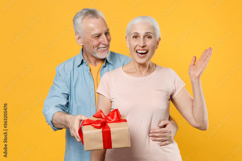 Excited elderly gray-haired couple woman man in casual clothes isolated on yellow background studio. St. Valentine's Day, Women's Day, birthday, holiday concept. Hold present box with gift ribbon bow. Excited elderly gray-haired couple woman man in casual clothes isolated on yellow background studio. St. Valentine's Day, Women's Day, birthday, holiday concept. Hold present box with gift ribbon bow.