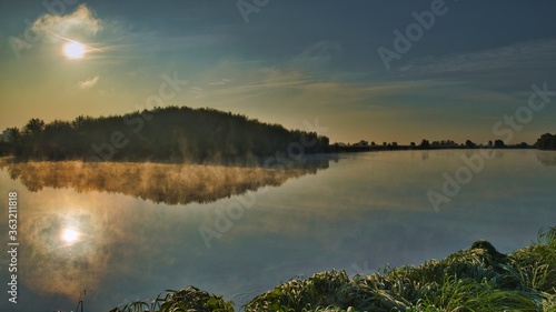 Reflection of the clear dawn sky, sun, forest in the smooth water of the river, foggy autumn morning © Kirill