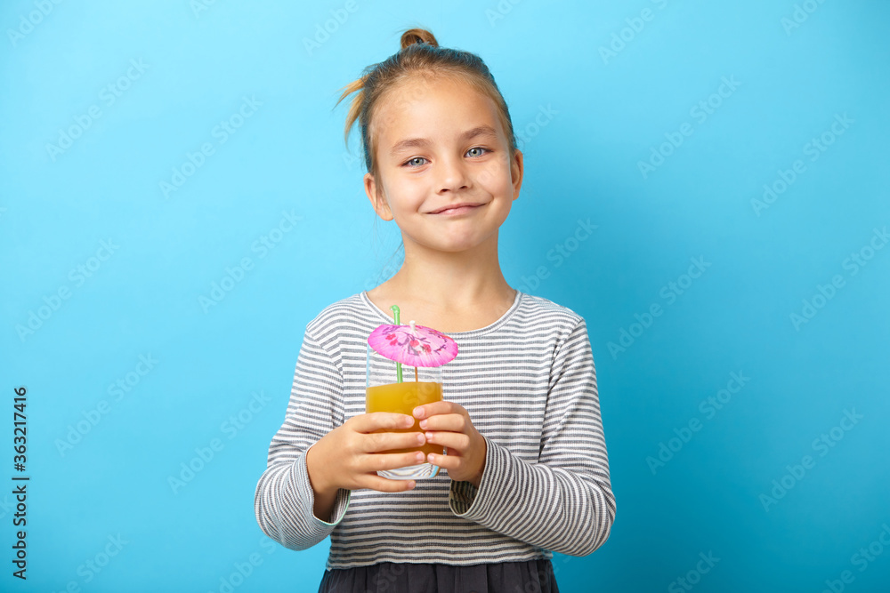 Smiling child girl drinks orange juice, has a good mood, stands against blue isolated background. Smiling child girl drinks orange juice, has a good mood, stands against blue isolated background.
