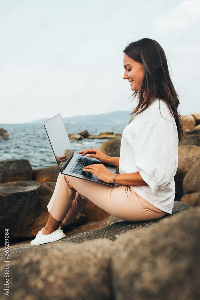Young successful, smiling girl working using a laptop, sitting on a rocky seashore. traveling, blogger, freelancer, content plan, work online