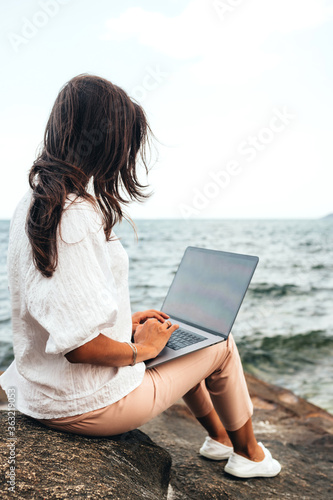 Young successful, smiling girl working using a laptop, sitting on a rocky seashore. traveling, blogger, freelancer, content plan, work online