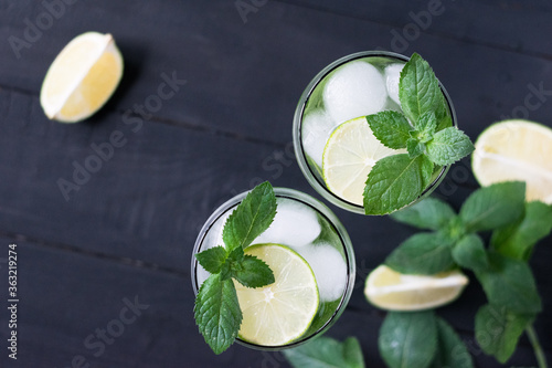 Two glasses of homemade citrus lemonade mojito with slices of lime, mint and ice on a dark wooden background.Top view.