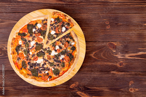 Pizza with cherry tomatoes, spinach, mozzarella, feta, kalamata olive and mushrooms, with a slice slightly removed on a wood platter which is on wooden rustic table, top view and copy space