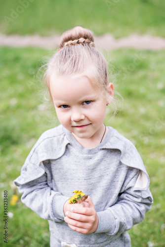 Little blond girl with blue eyes, hair arranged in a bun, wearing a gray dress, close-up portrait, looking into the camera, smiling, bored, playing on a lawn, sitting on the machine tires. Preschool 