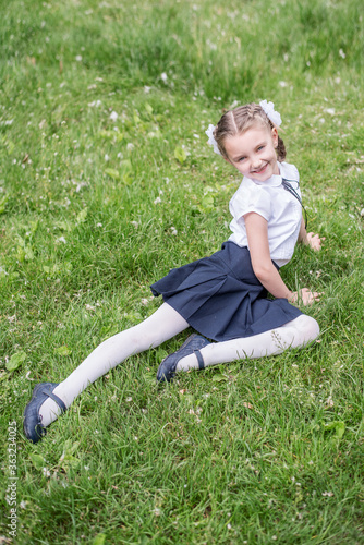 Little blonde schoolgirl in a school uniform with white bows on pigtails goes to first grade. Beginning the first academic trimester. Close-up portrait, looking at the camera, smiling toothless smile