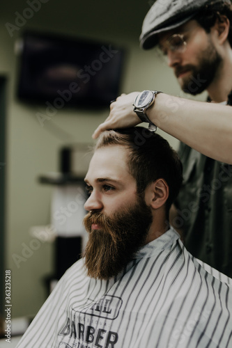 Great time at barbershop. Cheerful young bearded man getting haircut by hairdresser and reading newspaper while sitting in chair at barbershop