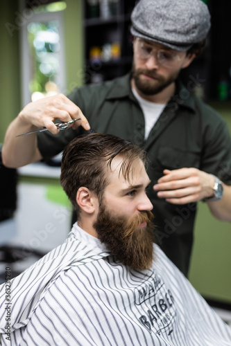 Side view of young bearded man getting beard haircut at hairdresser while sitting in chair at barbershop