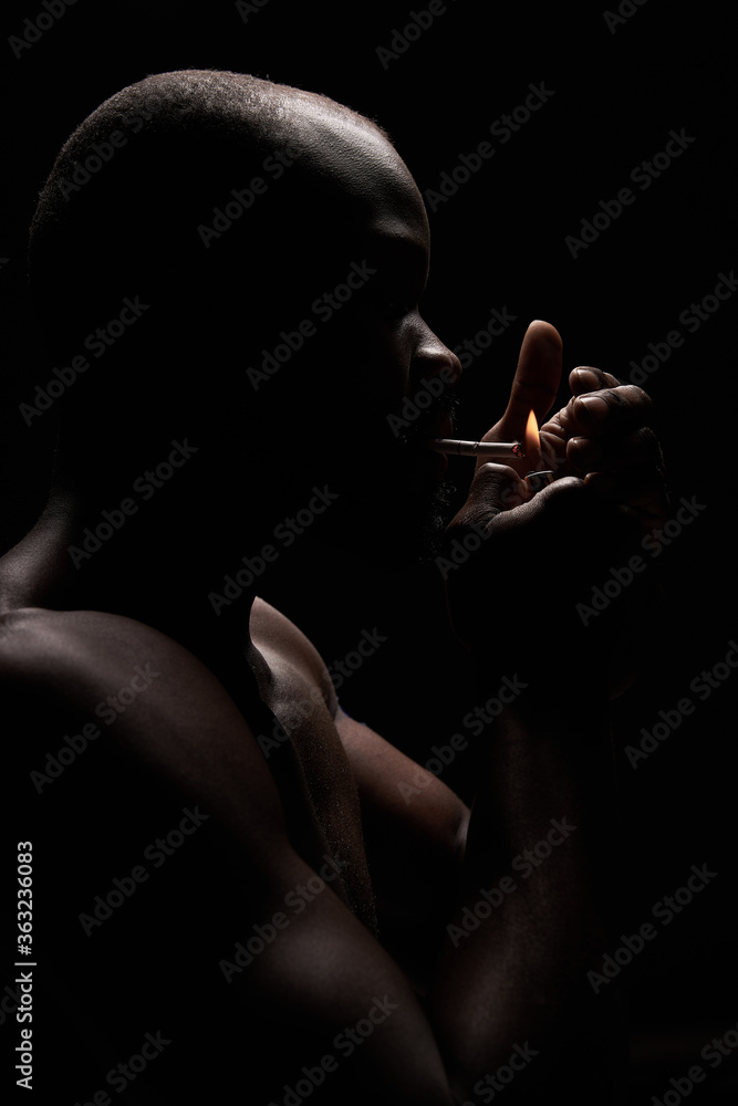 profile photo of a dark-skinned muscular handsome guy with a beard on a black background who lights a cigarette