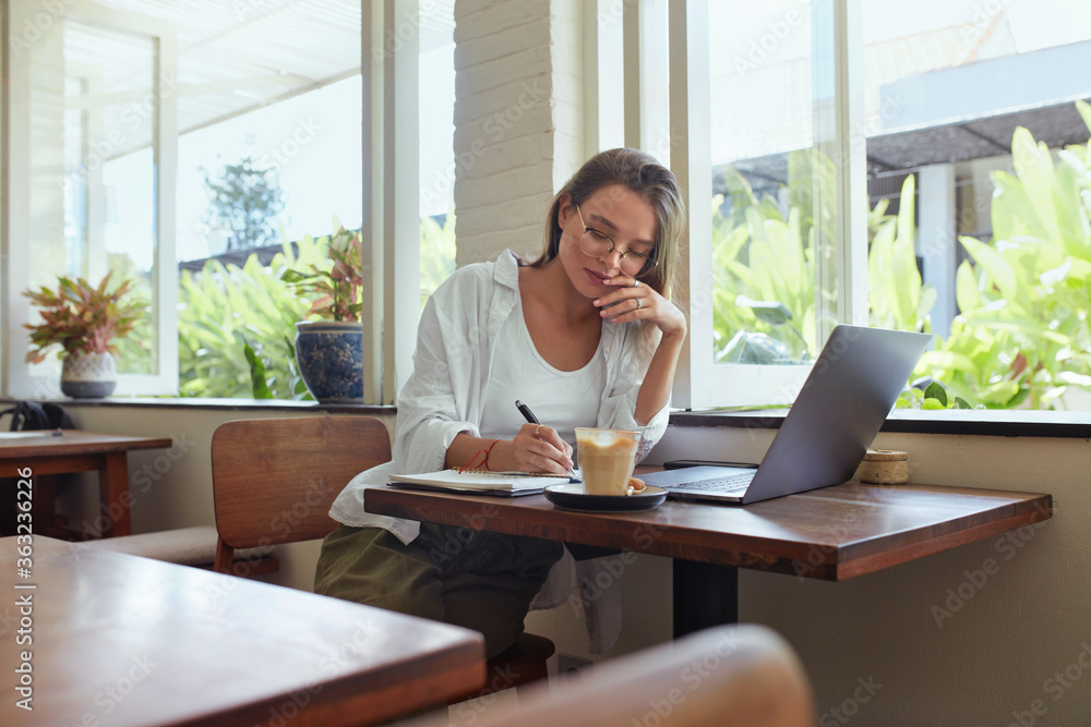 Girl. Student At Cafe With Laptop Writing Something. Beautiful Woman In Glasses Working Or Studying Remotely At Comfortable Workplace. Online Education Or Modern Creativity Job As Lifestyle. Girl. Student At Cafe With Laptop Writing Something. Beautiful Woman In Glasses Working Or Studying Remotely At Comfortable Workplace. Online Education Or Modern Creativity Job As Lifestyle.