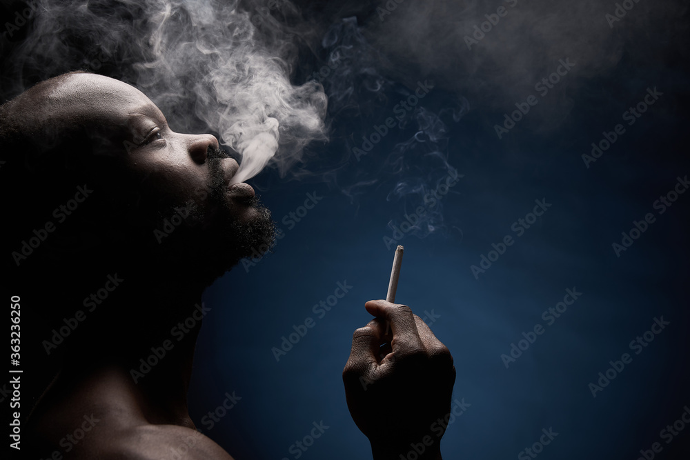 Fototapeta premium profile photo of a dark-skinned muscular handsome guy with a beard on a dark background who breathes out smoke from his mouth with a cigarette in his hand