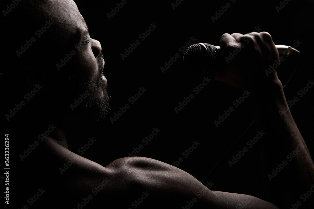 portrait of a dark-skinned muscular handsome guy with a beard on a black background who emotionally sings into a microphone