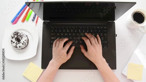 A young girl types on the laptop in the office, with sticky notes and a donut next to her. On a white table. Office overhead scene.